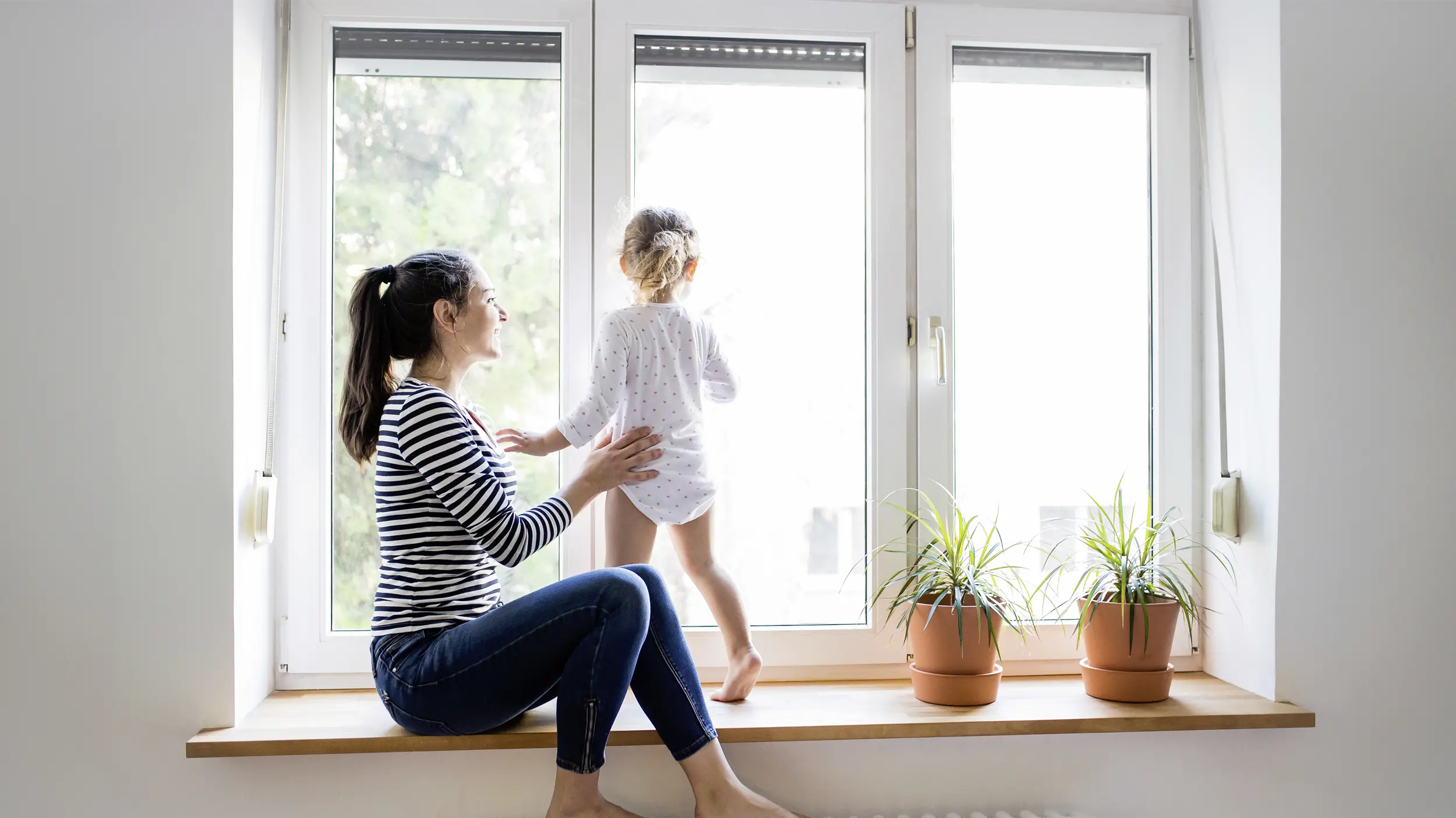 Eine Frau sitzt auf einem FEnstersims und hält ein Baby fest, das auf dem Sims steht undn aus dem Fenster schaut